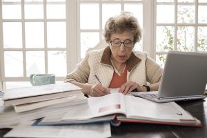 Elderly woman at a desk surrounded by papers, books, and a laptop.