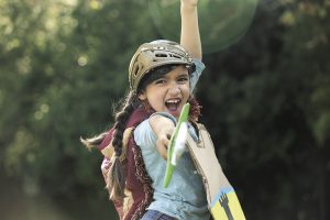 Child dressed up in homemade warrior outfit with cape, playfully pointing plastic sword at the camera.