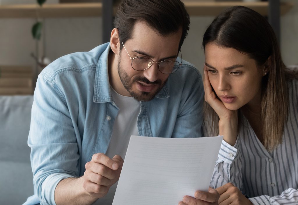 Serious pensive millennial 35s couple sit on sofa at home hold letter reading papers documents, learn details of contract, discuss agreement terms conditions, considering health care