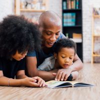 African American father reading a fairy tale fable story for kids at home. Happy family lying on the floor indoors